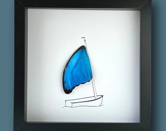 Pode incluir: Obra de arte emoldurada com uma asa de borboleta azul como vela de um veleiro. A obra é uma ilustração caprichosa com uma moldura preta, um fundo branco e um contorno simples de barco.
