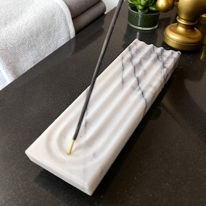 May include: A rectangular white marble incense holder featuring grey veining and a series of parallel, wavy grooves carved into the surface. A single thin black incense stick with a gold-coloured tip is positioned upright in the holder.