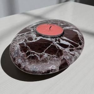 May include: A polished, oval-shaped candle holder crafted from dark brown marble with prominent white veining. The centre features a circular recessed area containing a small, round, coral-pink tea light candle with a single black wick.