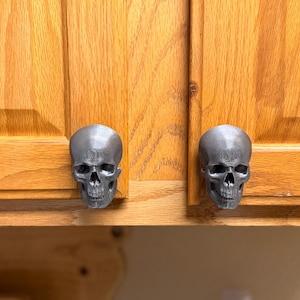 May include: Two metallic silver cabinet knobs shaped like human skulls. Each knob features detailed eye sockets, a nasal cavity, and a jawline, mounted onto the front of light brown wooden cabinet doors with a visible vertical wood grain pattern.