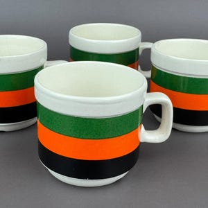Puede incluir: Un juego de cuatro tazas de cerámica con borde y asa blancos, decoradas con bandas horizontales de color verde, naranja y negro alrededor del cuerpo. Cada taza tiene una forma cilíndrica con un acabado liso y un asa lateral resistente.