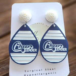 May include: A pair of blue and white teardrop-shaped earrings with the words "Cheer Leader" on them. The earrings are made of surgical steel and are hypoallergenic.