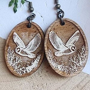 May include: Oval wooden earrings with a detailed white owl design in flight. The earrings have a rustic, natural aesthetic, with a carved owl and foliage on a brown background. The earrings hang from bronze-colored ear wires.