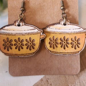 May include: A pair of wooden earrings shaped like yellow casserole dishes with floral cutouts. The earrings hang from bronze-colored fish hook ear wires and are displayed on a brown card.