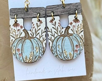Puede incluir: Pendientes colgantes de madera con un diseño de calabaza azul claro y detalles florales. Los pendientes tienen una parte superior marrón oscuro y ganchos dorados. El fondo es una tarjeta blanca con el texto "wicket goods".