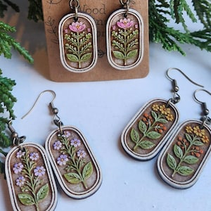 May include: Wooden earrings with floral designs. The earrings feature hand-painted flowers in pink, purple, and yellow, with green leaves. The earrings are rectangular with a light wood border. The text "Dawn's Wood" is visible on the tag.
