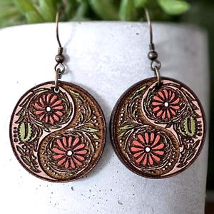 May include: A pair of round wooden earrings with a floral design. The earrings feature a brown background with a pink and green floral pattern. The earrings hang from bronze-colored hooks.