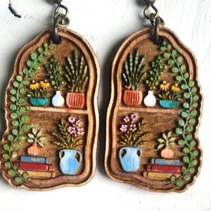 May include: Wooden earrings featuring a miniature bookshelf design with potted plants, flowers, and books. The earrings have a brown background with green, blue, yellow, and pink accents. The earrings hang from bronze-colored fish hook ear wires.