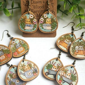 May include: Teardrop-shaped wooden earrings featuring colorful illustrations of cats in garden settings. The designs include cats in pots, wheelbarrows, and crates with flowers. The earrings have bronze-toned metal hooks and are displayed on a brown card labeled Dawn's Wood Earrings.