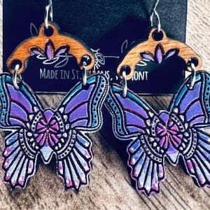 May include: A pair of butterfly earrings with a wooden top and a colorful butterfly design. The earrings have a silver hook and the text "Firefly & Fern" is visible on the black background.