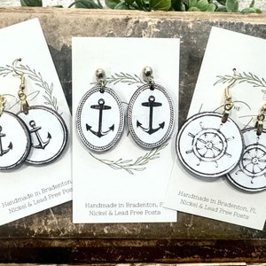 May include: Three pairs of earrings with nautical themes. The first pair features a black anchor on a white circle. The second pair features a black anchor on a white oval. The third pair features a black ship's wheel on a white circle. All earrings are handmade in Bradenton, Florida and are nickel and lead free.