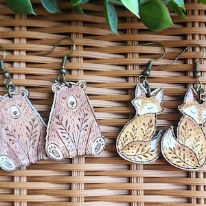 May include: Two pairs of dangle earrings featuring flat, illustrated animal charms. One pair depicts light brown bears with dark brown leaf patterns, and the other pair shows orange foxes with dark brown leaf patterns. Each charm hangs from a bronze-toned metal hook.