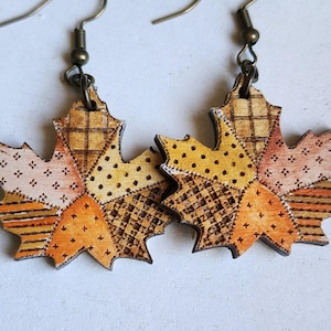 May include: A pair of maple leaf-shaped earrings made of wood, featuring a patchwork design with various patterns including dots, stripes, and grids in shades of orange, yellow, and brown, attached to dark metal fishhook ear wires.
