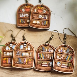 May include: Wooden earrings featuring a bookshelf design with various magical elements. Each earring showcases a miniature bookshelf filled with books, potion bottles, skulls, and other mystical items. The earrings have bronze-colored ear wires.