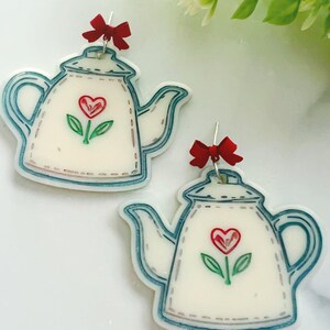 May include: A pair of white teapot earrings with a red heart and green stem design. The earrings have a blue outline and a small red bow at the top. The earrings are a whimsical accessory.