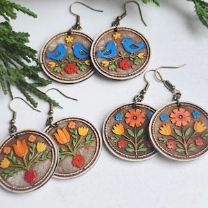 May include: Wooden earrings with a brown background and a white border. The earrings feature colorful designs of blue birds on a branch and flowers. The earrings have bronze-colored hooks.