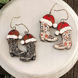 May include: Wooden earrings featuring cowboy boots with Santa hats. The boots are detailed with floral designs and a distressed finish. The Santa hats are red with white trim and a small white pom-pom. The earrings are on a white circular surface.