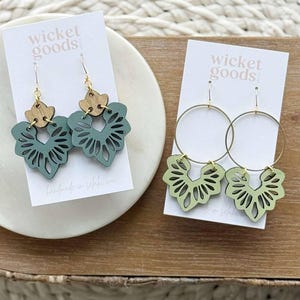 Puede incluir: Dos pares de pendientes en tarjetas blancas. Uno presenta un diseño calado con láser en verde salvia con ganchos dorados y detalles de madera. El otro par tiene un diseño similar en verde claro, con aros dorados. Las tarjetas dicen "wicket goods".