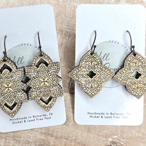 May include: Two pairs of laser-cut wooden earrings with intricate floral and geometric designs. The earrings are displayed on white cards, with black ear wires. The text "Handmade in Bulverde, TX" is visible on the cards.