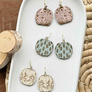 May include: Wooden pumpkin earrings in three different color schemes: pink with brown floral details, light blue with brown floral details, and cream with brown leaf details. The earrings are displayed on a white tray with a wooden slice and woven basket in the background.