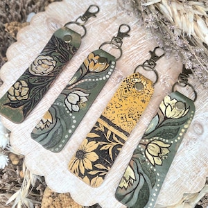 May include: Four leather keychains with floral designs. The keychains have a green base color with hand-painted white and gold flowers. Each keychain has a metal clasp and rivet for attachment. The keychains are displayed on a wooden surface.
