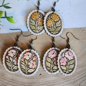 May include: Six pairs of wooden earrings with floral designs. Two pairs feature yellow flowers, while the other four pairs display pink flowers. Each earring has a brown oval center with a white scalloped edge and a metal hook.