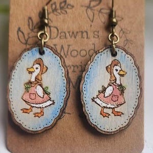 May include: Wooden earrings featuring a hand-painted illustration of a duck wearing a bonnet and dress. The duck is set against a light blue background with a scalloped border. The earrings hang from bronze-colored ear wires and are attached to a brown card.