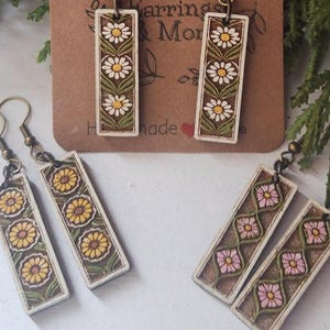 Puede incluir: Pendientes de madera con diseños florales. Los pendientes presentan colgantes rectangulares con diseños de margaritas, girasoles y flores rosas. La tarjeta de fondo dice "Wood Earrings & More" y "handmade".