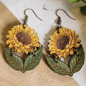 Pode incluir: Um par de brincos de girassol de madeira. Cada brinco apresenta um girassol amarelo com um centro castanho e folhas verdes. Os brincos penduram-se em ganchos de cor bronze.