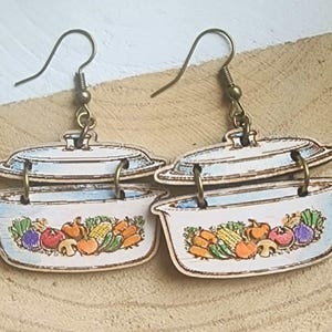 May include: Wooden earrings in the shape of casserole dishes. The earrings show a white dish with a blue rim and lid, decorated with colourful vegetable illustrations. They hang from bronze-coloured ear wires.