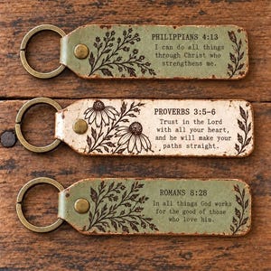 May include: Three rectangular keychains with metal rings and brass rivets. The top is sage green with Philippians 4:13, the middle is cream with Proverbs 3:5-6, and the bottom is sage green with Romans 8:28. Each features black floral illustrations.
