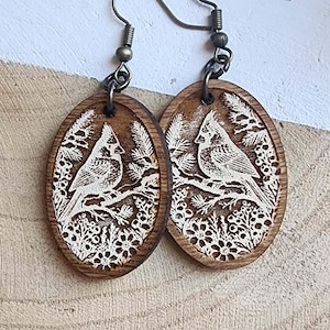 May include: A pair of wooden oval earrings with a detailed laser-cut design. Each earring features a white cardinal bird perched on a branch, surrounded by foliage. The earrings have bronze-colored ear wires.