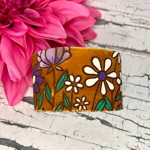 May include: A brown fabric bracelet with a floral pattern featuring white, purple, and green flowers.