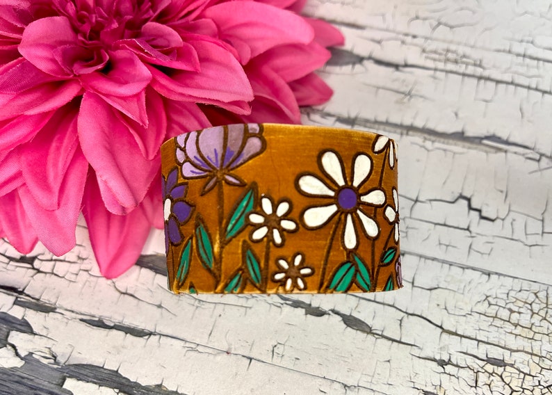 May include: A brown fabric bracelet with a floral pattern featuring white, purple, and green flowers.