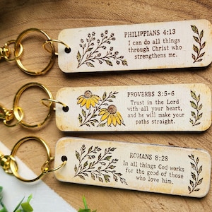 May include: Three rectangular wooden keychains with gold-tone metal lobster clasps. Each keychain features a unique Bible verse, black printed text, and botanical illustrations. One keychain includes yellow flower accents alongside the text.