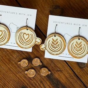 Könnte beinhalten: Zwei Paar hölzerne Kaffeetassen-Ohrringe mit Latte-Art-Motiven, eines mit Herz- und das andere mit Farnmuster. Die Ohrringe sind auf weißen Karten mit der Aufschrift Adventures With Jen befestigt, zusammen mit vier kleinen passenden Kaffeetassen-Anhängern.