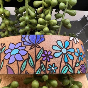 May include: A brown leather band with a floral design. The flowers are blue, purple, and teal. The band is decorated with a variety of flowers and leaves.
