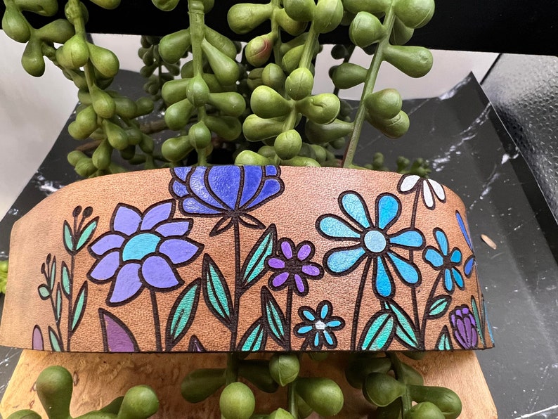 May include: A brown leather band with a floral design. The flowers are blue, purple, and teal. The band is decorated with a variety of flowers and leaves.