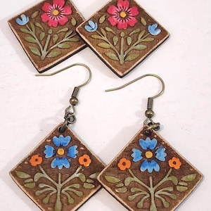 May include: Hand-painted wooden earrings with a floral design. The earrings feature a square shape with a brown background and colorful flowers in pink, blue, and orange. The earrings have brass-colored ear wires.
