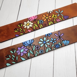 May include: Two brown leather bookmarks with colorful floral designs. The top bookmark features pink, yellow, orange, and green flowers. The bottom bookmark features blue, purple, and green flowers.
