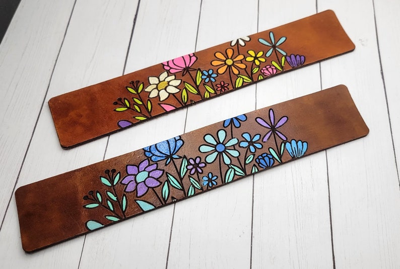 May include: Two brown leather bookmarks with colorful floral designs. The top bookmark features pink, yellow, orange, and green flowers. The bottom bookmark features blue, purple, and green flowers.