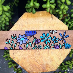May include: A wooden strip with a hand-painted design of blue, purple, and teal flowers. The flowers are outlined in black and have a whimsical, folk art style.