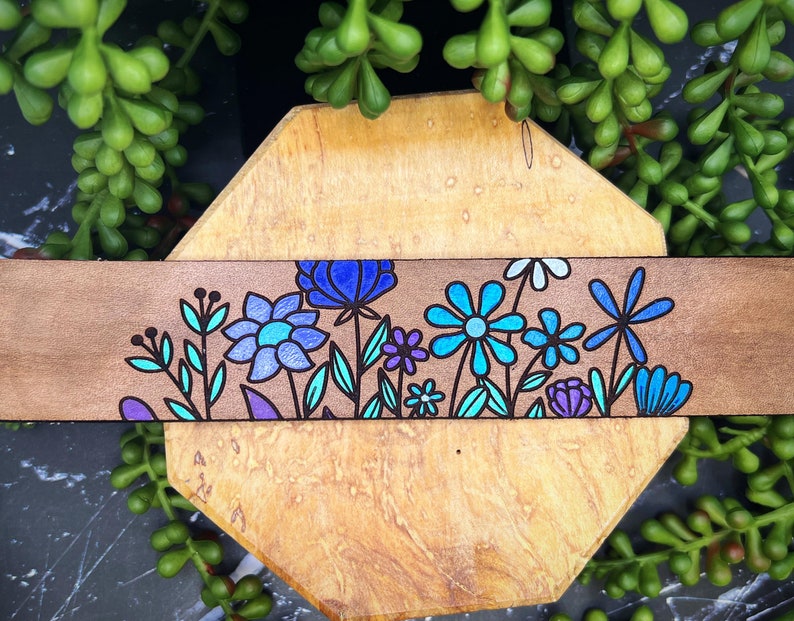 May include: A wooden strip with a hand-painted design of blue, purple, and teal flowers. The flowers are outlined in black and have a whimsical, folk art style.