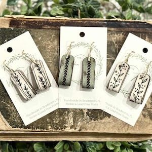Op de afbeelding: Handgemaakte oorbellen met botanische ontwerpen. De oorbellen zijn rechthoekig met een crèmekleurige achtergrond en zwarte bladerpatronen. Eén paar is olijfgroen met een zwart chevron-ontwerp. De oorbellen worden op kaarten gepresenteerd.