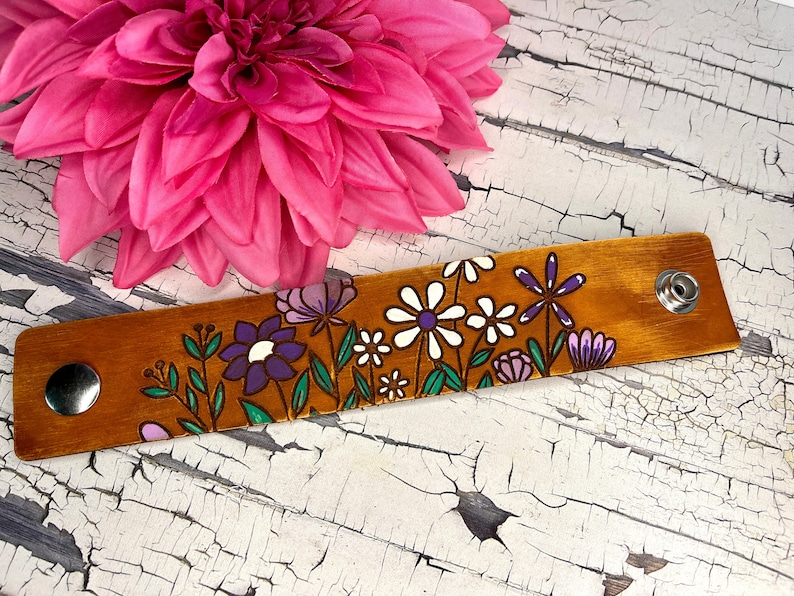 May include: A brown leather bracelet with a floral design painted on it. The flowers are purple, white, and green. The bracelet has two silver snaps.