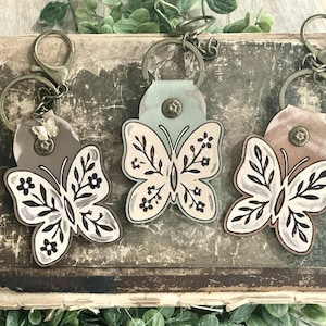 May include: Three butterfly keychains with antique brass hardware. Each butterfly has a unique design with black and white details. The keychains are attached to a leather backing in shades of brown and green.