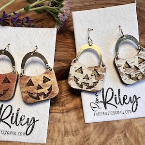May include: Wooden Halloween earrings in the shape of jack-o'-lanterns. The earrings have a gold-colored handle and a carved face. The earrings are on white cards with the text "Penny Riley" and an email address.