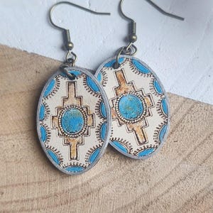 May include: Oval-shaped earrings with a Southwestern design. The earrings feature a cream-colored background with a turquoise center stone and accents. The design includes a cross-like shape in a tan color, surrounded by turquoise dots. The earrings have bronze-colored hooks.