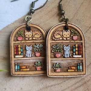 May include: Wooden earrings featuring a bookshelf design with cats, plants, and books. The earrings have a light brown background with detailed illustrations of the items on the shelves. The earrings hang from bronze-colored ear wires.