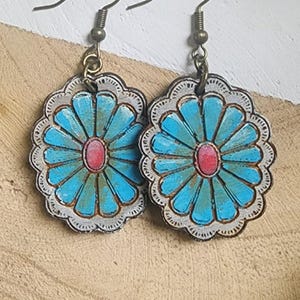 May include: A pair of turquoise and red flower-shaped earrings with a distressed, vintage look. The earrings have a scalloped edge and are suspended from bronze-colored ear wires. The floral design features a central red oval surrounded by turquoise petals.